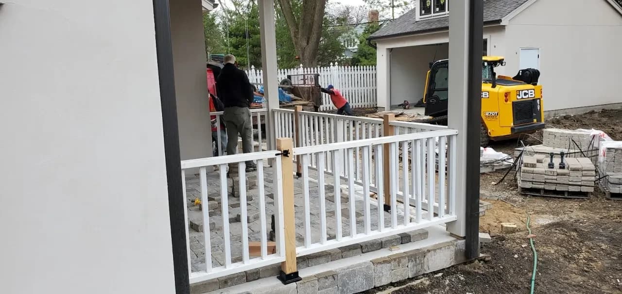 A crew laying pavers and building a porch rail, with a loader behind